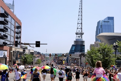 A vibrant street scene with a large crowd of people participating in a parade. Many are carrying rainbow umbrellas and wearing colorful clothing, indicating a celebration of diversity and inclusion. Tall buildings line the street, with a distinctive modern skyscraper visible in the background. Trees and traffic lights are also present, adding to the lively urban atmosphere.