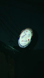 A round sign for a Mexican taqueria featuring various elements, including the name 'La Tapatia'. There are several logos and symbols, such as a taco graphic and flags of multiple countries. The sign is illuminated in a dark setting with visible electrical wires in the background.