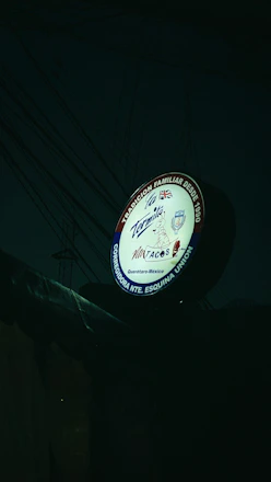 A round sign for a Mexican taqueria featuring various elements, including the name 'La Tapatia'. There are several logos and symbols, such as a taco graphic and flags of multiple countries. The sign is illuminated in a dark setting with visible electrical wires in the background.