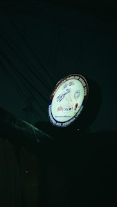 A round sign for a Mexican taqueria featuring various elements, including the name 'La Tapatia'. There are several logos and symbols, such as a taco graphic and flags of multiple countries. The sign is illuminated in a dark setting with visible electrical wires in the background.