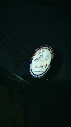 A round sign for a Mexican taqueria featuring various elements, including the name 'La Tapatia'. There are several logos and symbols, such as a taco graphic and flags of multiple countries. The sign is illuminated in a dark setting with visible electrical wires in the background.