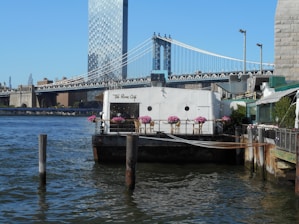 A riverside café with outdoor seating adorned with pink flowers overlooks a body of water. In the background, a large bridge spans across the scene, and a tall, mirrored skyscraper rises into the blue sky. The café appears welcoming, with a few tables and chairs arranged neatly on the deck.