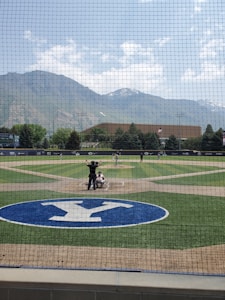 A baseball field is visible with a game in progress. Players are positioned on the field, and an umpire stands behind the catcher. The field has a large blue and white logo near home plate and a background of mountains covered with scattered clouds.