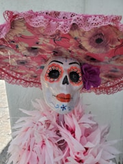 A decorative skeleton head with colorful face paint, featuring orange eye accents, orange lips, and floral embellishments. It is wearing a large, circular hat covered in floral patterns and pink lace trimming. Around its neck is a draped arrangement of pink paper-like material resembling feathers or flowers.
