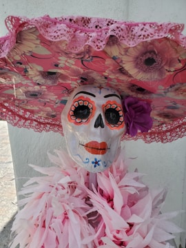 A decorative skeleton head with colorful face paint, featuring orange eye accents, orange lips, and floral embellishments. It is wearing a large, circular hat covered in floral patterns and pink lace trimming. Around its neck is a draped arrangement of pink paper-like material resembling feathers or flowers.