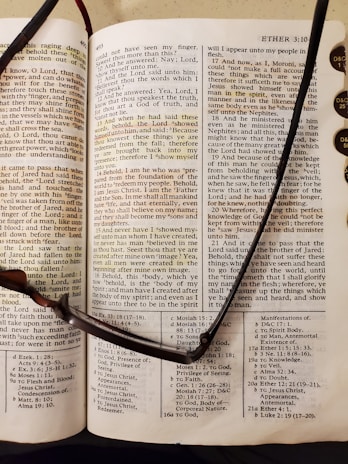 An open book with text details from a religious scripture. The page appears to be from the Book of Mormon, showcasing chapter and verse numbers. A pair of reading glasses rests across the open pages, indicating someone may have been reading. Nearby, tabs with lettering are visible, likely for easy navigation between sections.