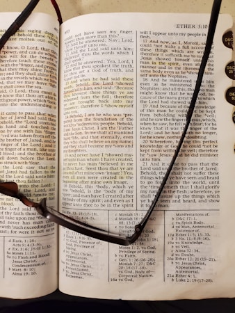 An open book with text details from a religious scripture. The page appears to be from the Book of Mormon, showcasing chapter and verse numbers. A pair of reading glasses rests across the open pages, indicating someone may have been reading. Nearby, tabs with lettering are visible, likely for easy navigation between sections.
