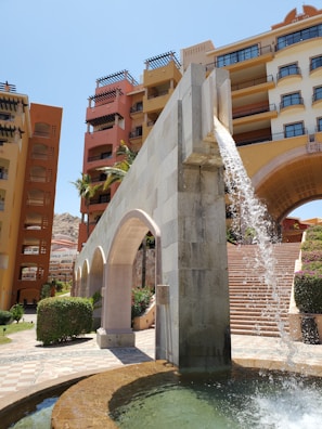 A cascading water feature descends from a stone aqueduct-like structure into a small pool, set against a backdrop of colorful, multi-story buildings with balconies. Palm trees and manicured bushes add to the lush ambiance, while a set of stairs leads upward, suggesting an inviting pathway.
