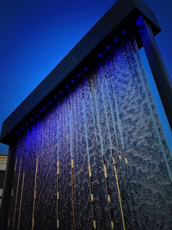 A modern architectural installation featuring a large, vertical panel with cascading water illuminated by blue lights. The panel appears to have a textured surface, and the lighting creates a vibrant contrast against the dark background.