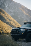 A rugged Toyota truck is parked on a gravel path, surrounded by dense forested hills and a towering mountain in the background. The sunlight casts a warm glow over the landscape, highlighting the contours of the trees and mountain.