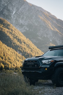 A rugged Toyota truck is parked on a gravel path, surrounded by dense forested hills and a towering mountain in the background. The sunlight casts a warm glow over the landscape, highlighting the contours of the trees and mountain.