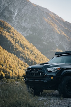 A rugged Toyota truck is parked on a gravel path, surrounded by dense forested hills and a towering mountain in the background. The sunlight casts a warm glow over the landscape, highlighting the contours of the trees and mountain.