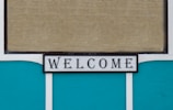 A decorative sign displaying the word 'WELCOME' in bold, black letters on a white background with a thin black border. The background consists of teal panels and a beige, textured surface, possibly a window blind.