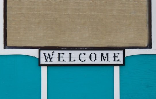 A decorative sign displaying the word 'WELCOME' in bold, black letters on a white background with a thin black border. The background consists of teal panels and a beige, textured surface, possibly a window blind.