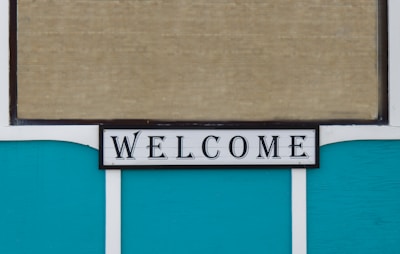 A decorative sign displaying the word 'WELCOME' in bold, black letters on a white background with a thin black border. The background consists of teal panels and a beige, textured surface, possibly a window blind.