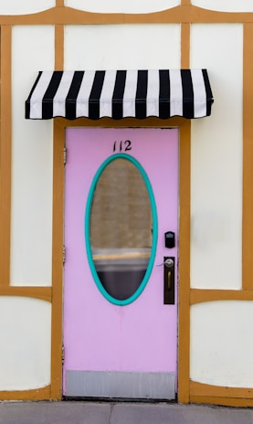 A pastel pink door with an oval-shaped window and turquoise trim is set into a cream and wooden frame wall. Above the door hangs a black and white striped awning. The door has a silver handle and an electronic lock. The number 112 is displayed above the window.