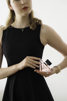 A person in a sleeveless black dress holds a bottle of Prada perfume. The individual wears a gold watch and a delicate chain necklace with a cross pendant. The background is neutral with soft lighting, focusing attention on the person and the perfume bottle.