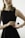 A person in a sleeveless black dress holds a bottle of Prada perfume. The individual wears a gold watch and a delicate chain necklace with a cross pendant. The background is neutral with soft lighting, focusing attention on the person and the perfume bottle.