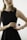 A person in a sleeveless black dress holds a bottle of Prada perfume. The individual wears a gold watch and a delicate chain necklace with a cross pendant. The background is neutral with soft lighting, focusing attention on the person and the perfume bottle.