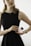 A person in a sleeveless black dress holds a bottle of Prada perfume. The individual wears a gold watch and a delicate chain necklace with a cross pendant. The background is neutral with soft lighting, focusing attention on the person and the perfume bottle.