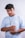 A person wearing a light blue t-shirt with the text 'To Love is to Forgive' stands against a white textured background. They have a short haircut and a focused expression. A black cross necklace and a wristwatch are visible, along with a bracelet on one wrist.