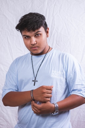 A person wearing a light blue t-shirt with the text 'To Love is to Forgive' stands against a white textured background. They have a short haircut and a focused expression. A black cross necklace and a wristwatch are visible, along with a bracelet on one wrist.
