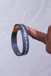 A hand holding a black lanyard or wristband between two fingers. The band has white text with the words 'external' and 'internal' printed on it. The background is a plain, light-colored backdrop.