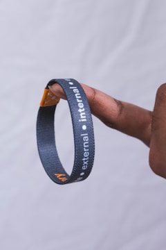 A hand holding a black lanyard or wristband between two fingers. The band has white text with the words 'external' and 'internal' printed on it. The background is a plain, light-colored backdrop.