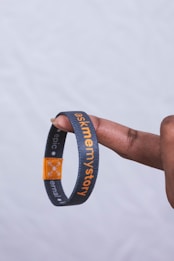 A black wristband with the phrase 'ask me my story' in orange text is placed on a finger. The band also has a small orange section with white symbols and additional text.