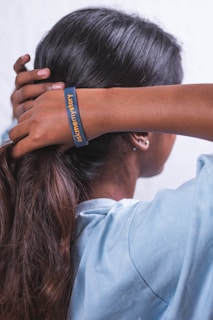 A person with long hair is seen from the back, wearing a light blue shirt. Their hands are adjusting their hair, and a bracelet on one wrist reads 'ask me my story'.