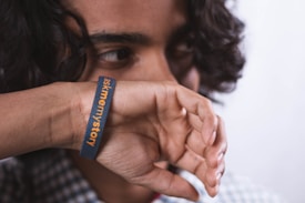 A person with curly hair covers their mouth with their hand, wearing a wristband that reads 'ask me my story' in orange and white text. The person has a thoughtful expression and is partially obscured by their hand.