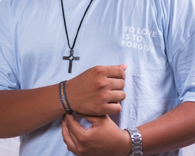 A person is wearing a light blue shirt with the words 'TO LOVE IS TO FORGIVE' printed on it. They have a black necklace with a cross pendant around their neck. A wristband on their right hand reads 'ask me my story,' and they wear a metallic watch on their left wrist.