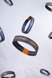 Several dark wristbands with orange and white text float against a light, textured background. The text includes the phrases 'askmemystory' and words like 'external,' 'internal,' and 'epic'. An orange square graphic is also visible on each band.