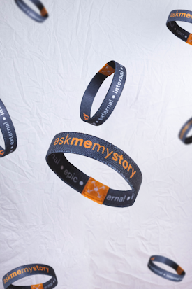 Several dark wristbands with orange and white text float against a light, textured background. The text includes the phrases 'askmemystory' and words like 'external,' 'internal,' and 'epic'. An orange square graphic is also visible on each band.