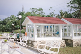 Several white gazebos with red roofs are situated in a garden setting, surrounded by lush green trees. The gazebos have large windows and are elevated on stone foundations. White benches and lampposts line a paved walkway leading to the structures.