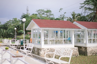 Several white gazebos with red roofs are situated in a garden setting, surrounded by lush green trees. The gazebos have large windows and are elevated on stone foundations. White benches and lampposts line a paved walkway leading to the structures.