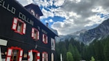 A black building with red and white window frames stands in the foreground. The structure is surrounded by tall pine trees and beneath a partly cloudy sky. In the background, rocky mountains are visible.