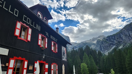 A black building with red and white window frames stands in the foreground. The structure is surrounded by tall pine trees and beneath a partly cloudy sky. In the background, rocky mountains are visible.