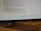 A close-up of an open book page featuring religious text in a serif font. The page is resting on a wooden surface, showcasing partial lines of text and a visible border of the book binding.