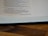 A close-up of an open book page featuring religious text in a serif font. The page is resting on a wooden surface, showcasing partial lines of text and a visible border of the book binding.
