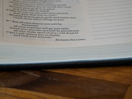A close-up of an open book page featuring religious text in a serif font. The page is resting on a wooden surface, showcasing partial lines of text and a visible border of the book binding.