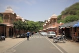 A street scene features traditional Indian architecture with ornate facades and domes on either side. Several people walk along the street, and motorbikes and cars are parked along the sides. Trees and greenery are visible, adding a natural element to the otherwise bustling street. Stalls with signs and bikes lined up contribute to the lively atmosphere.
