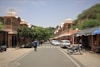 A street scene features traditional Indian architecture with ornate facades and domes on either side. Several people walk along the street, and motorbikes and cars are parked along the sides. Trees and greenery are visible, adding a natural element to the otherwise bustling street. Stalls with signs and bikes lined up contribute to the lively atmosphere.