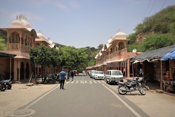 A street scene features traditional Indian architecture with ornate facades and domes on either side. Several people walk along the street, and motorbikes and cars are parked along the sides. Trees and greenery are visible, adding a natural element to the otherwise bustling street. Stalls with signs and bikes lined up contribute to the lively atmosphere.
