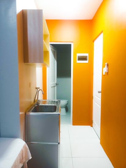 A small, brightly lit room with vivid orange walls and a white ceiling. There is a stainless steel sink with a faucet on the left, adjacent to a wall-mounted wooden cabinet. The room leads to a white door that opens into a space with a visible toilet. The floor is tiled with large white squares.