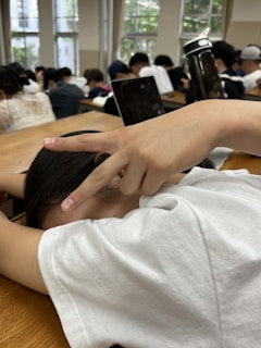 A person wearing a white shirt is sitting at a wooden desk with their head down, resting. Their hand is raised, making a peace sign. In the background, there are several other people seated, focused, with a large water bottle and a laptop visible. The room has large windows with natural light coming in.