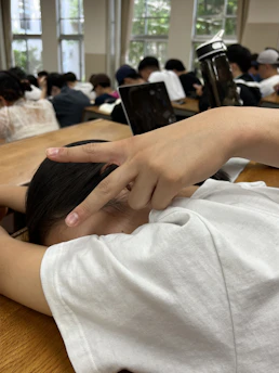 A person wearing a white shirt is sitting at a wooden desk with their head down, resting. Their hand is raised, making a peace sign. In the background, there are several other people seated, focused, with a large water bottle and a laptop visible. The room has large windows with natural light coming in.