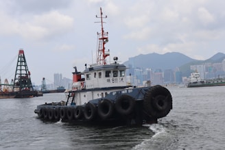 A harbor scene with a tugboat prominently in the foreground, featuring numerous tires along its sides. In the background, an urban skyline with high-rise buildings and mountains is visible, alongside other ships and a large crane structure on the water.