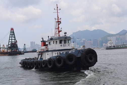 A harbor scene with a tugboat prominently in the foreground, featuring numerous tires along its sides. In the background, an urban skyline with high-rise buildings and mountains is visible, alongside other ships and a large crane structure on the water.