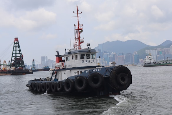 A harbor scene with a tugboat prominently in the foreground, featuring numerous tires along its sides. In the background, an urban skyline with high-rise buildings and mountains is visible, alongside other ships and a large crane structure on the water.